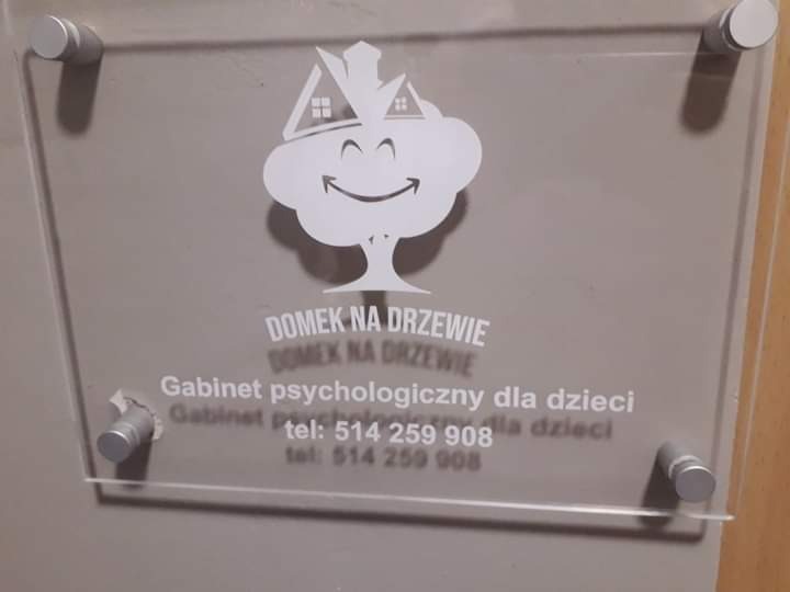 Tabliczka z logo i napisem 'Domek na Drzewie, Gabinet psychologiczny dla dzieci' przymocowana do ściany za pomocą srebrnych śrub dystansowych.
