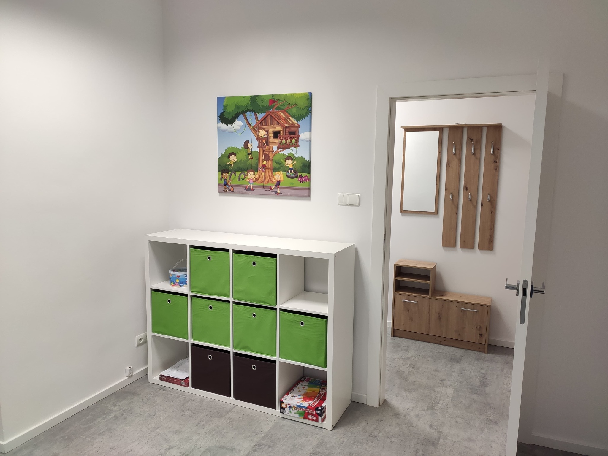 Jasne wnętrze gabinetu z białymi ścianami, regałem z zielonymi i brązowymi pojemnikami, obrazkiem dziecięcego domku na drzewie i widokiem na przedpokój z szafką na buty i wieszakami.