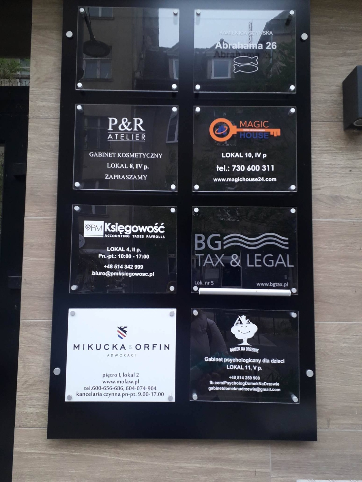 Tablica informacyjna z szyldami różnych firm, w tym gabinetu psychologicznego dla dzieci 'Domek na Drzewie' z symbolem drzewa i uśmiechniętej buzi, oraz innych firm z branży kosmetycznej, księgowej...