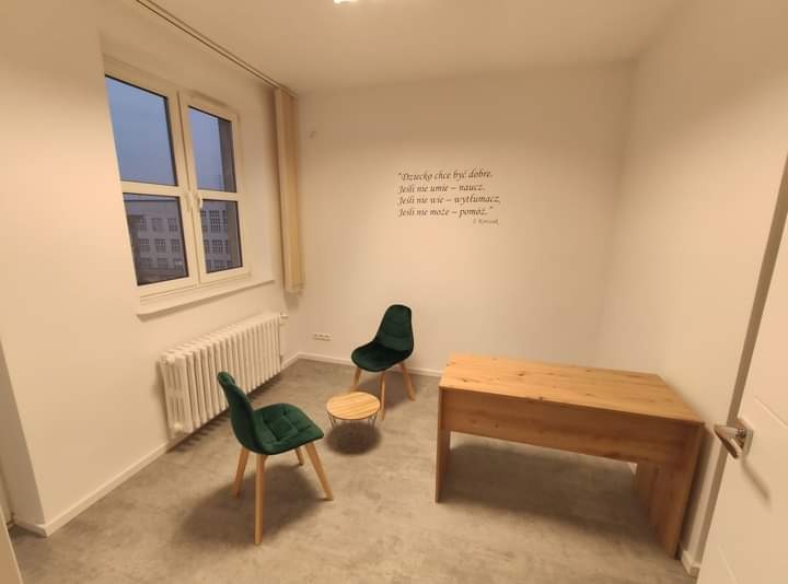 Jasne wnętrze gabinetu z drewnianym biurkiem, dwoma zielonymi krzesłami, małym okrągłym stolikiem i cytatem Janusza Korczaka na ścianie.