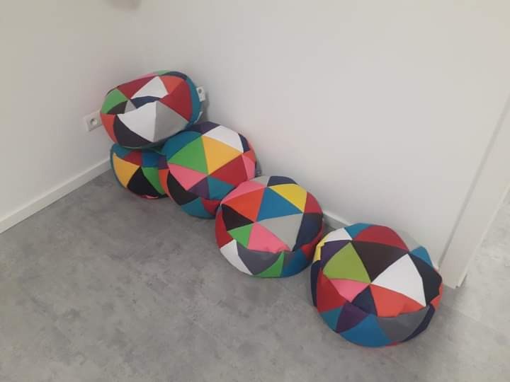 Pięć kolorowych, patchworkowych piłek relaksacyjnych ułożonych w rogu pokoju na szarej podłodze, przy białej ścianie z widocznym gniazdkiem elektrycznym.
