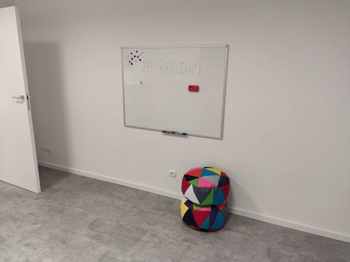 Jasne, minimalistyczne wnętrze gabinetu z białą tablicą z napisem 'Zapraszam', kolorowa pufa w rogu i otwarte białe drzwi.