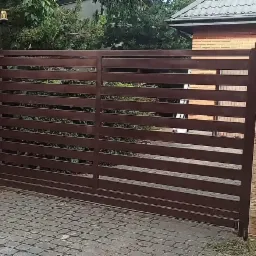 Zamykana, przesuwna brama wjazdowa z poziomych, brązowych profili, osadzona pomiędzy dwoma filarami z okładziną imitującą piaskowiec, na tle domu z cegły i zieleni.