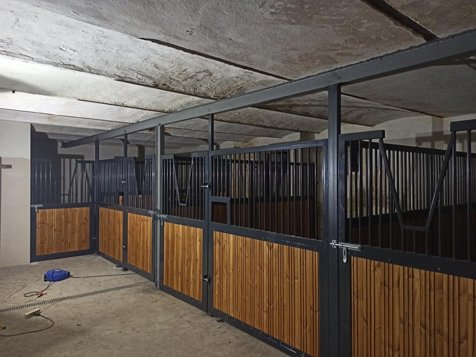 Wnętrze stajni z nowymi boksami dla koni, z drewnianymi panelami pionowymi i metalowymi kratami, surowy betonowy sufit, widoczny niebieski przedłużacz na podłodze.