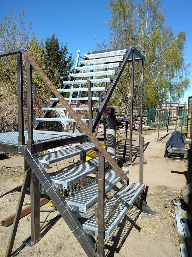 Szkielet metalowych schodów zewnętrznych z kratowymi stopniami, spawane elementy konstrukcyjne, widoczny sprzęt spawalniczy w tle, surowy stan przed malowaniem.