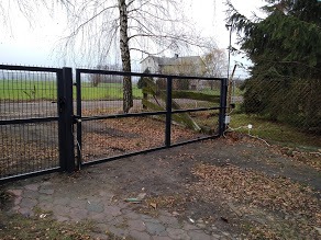 Czarna, metalowa brama wjazdowa z siatkowym wypełnieniem na posesję, widok z bliska na mechanizm otwierający, w tle zarośnięte pole i budynek gospodarczy.