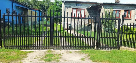 Czarna, metalowa brama wjazdowa i fragment ogrodzenia posesji z widocznymi dwoma budynkami w tle.