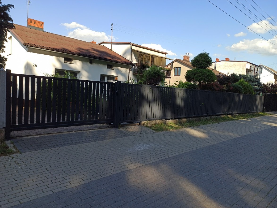 Ciemna, metalowa brama wjazdowa przesuwna z pionowych paneli, z numerem domu '26' widocznym na budynku w tle, na tle domów jednorodzinnych w słoneczny dzień.