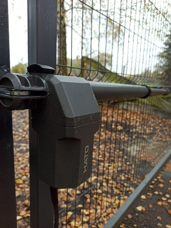 Szary automat do bramy z logo HATO, zamontowany na metalowej siatce ogrodzeniowej, z opadłymi liśćmi w tle.