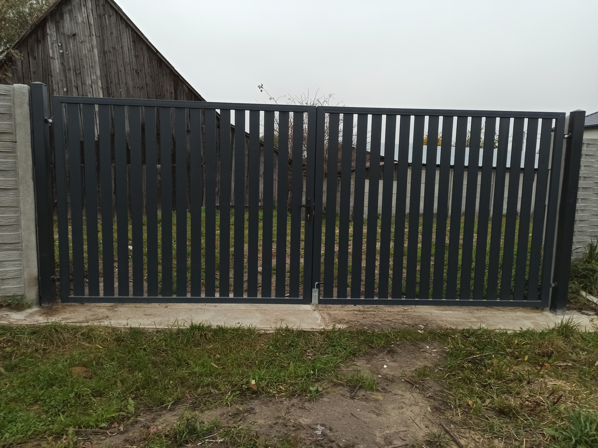 Nowoczesna brama wjazdowa z pionowych, grafitowych profili, z widocznym zamkiem, osadzona na betonowej podstawie i słupkach imitujących kamień, na tle pochmurnego nieba i drewnianego domu.