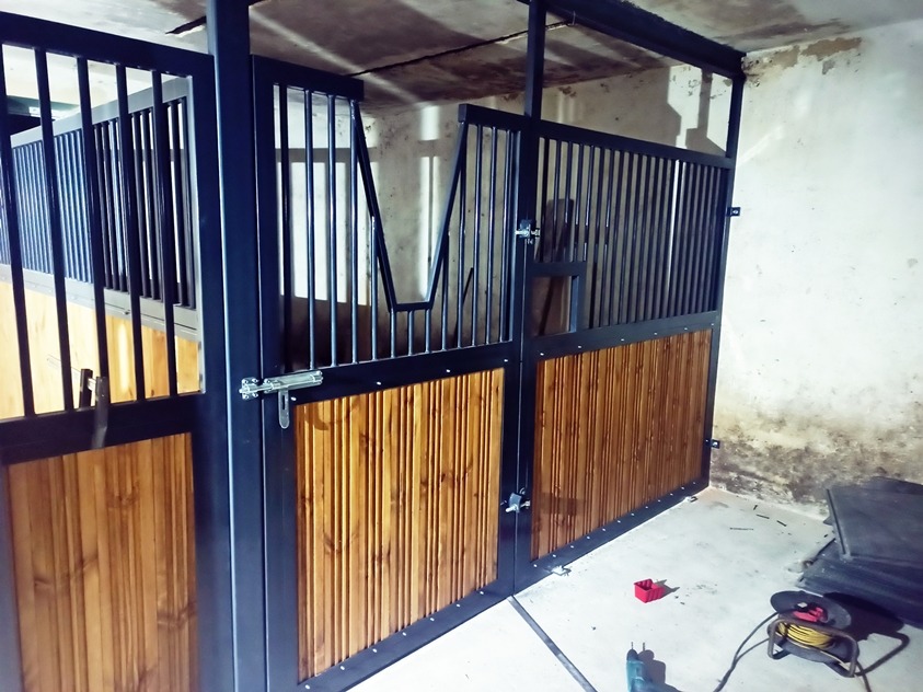 Konstrukcja boksów dla koni z metalową ramą i drewnianym wypełnieniem, widoczne zasuwy i narzędzia na podłodze.