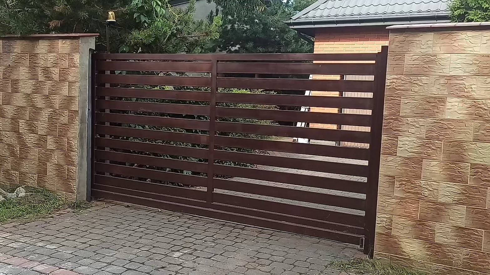Zamykana, przesuwna brama wjazdowa z poziomych, brązowych profili, osadzona pomiędzy dwoma filarami z okładziną imitującą piaskowiec, na tle domu z cegły i zieleni.