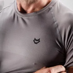 Mężczyzna w szarej, dopasowanej koszulce z krótkim rękawem i małym, czarnym logo na piersi, prezentujący ją w zbliżeniu.