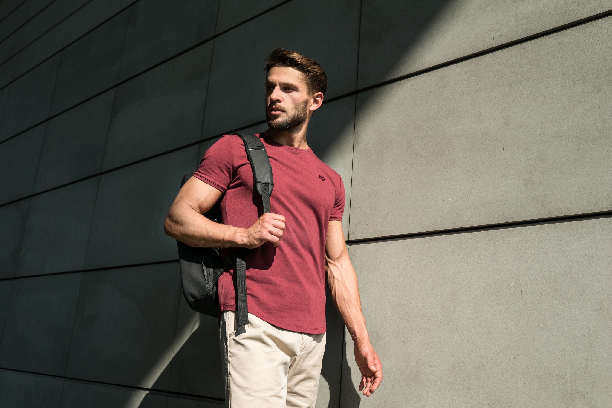 Mężczyzna w bordowej koszulce i beżowych spodniach z czarnym plecakiem, pozujący na tle szarej, nowoczesnej ściany z wyraźnymi liniami, oświetlony mocnym światłem słonecznym.