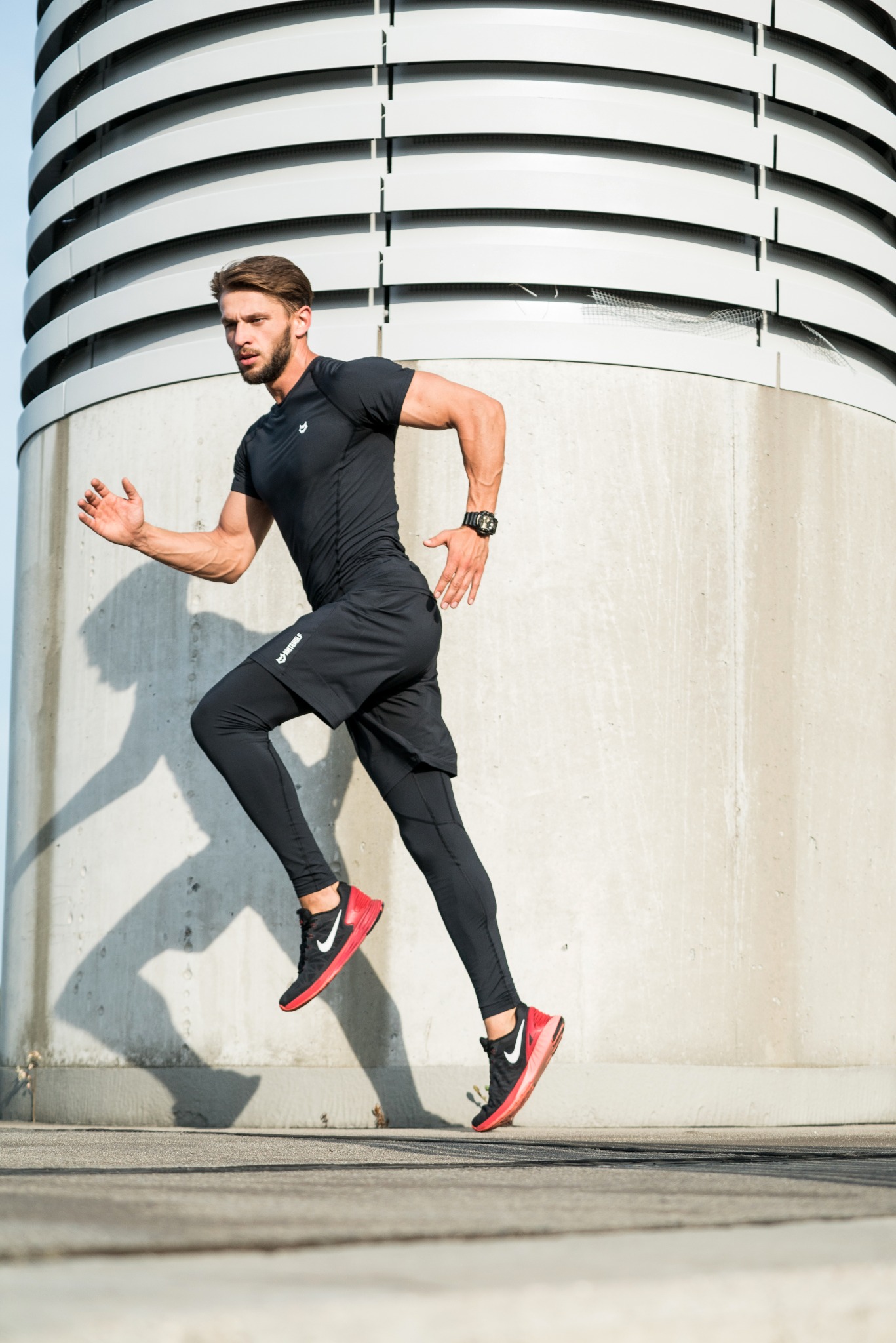 Mężczyzna w czarnym sportowym stroju i czerwono-czarnych butach do biegania, w dynamicznym ruchu, biegnie na tle betonowej konstrukcji z poziomymi żaluzjami.