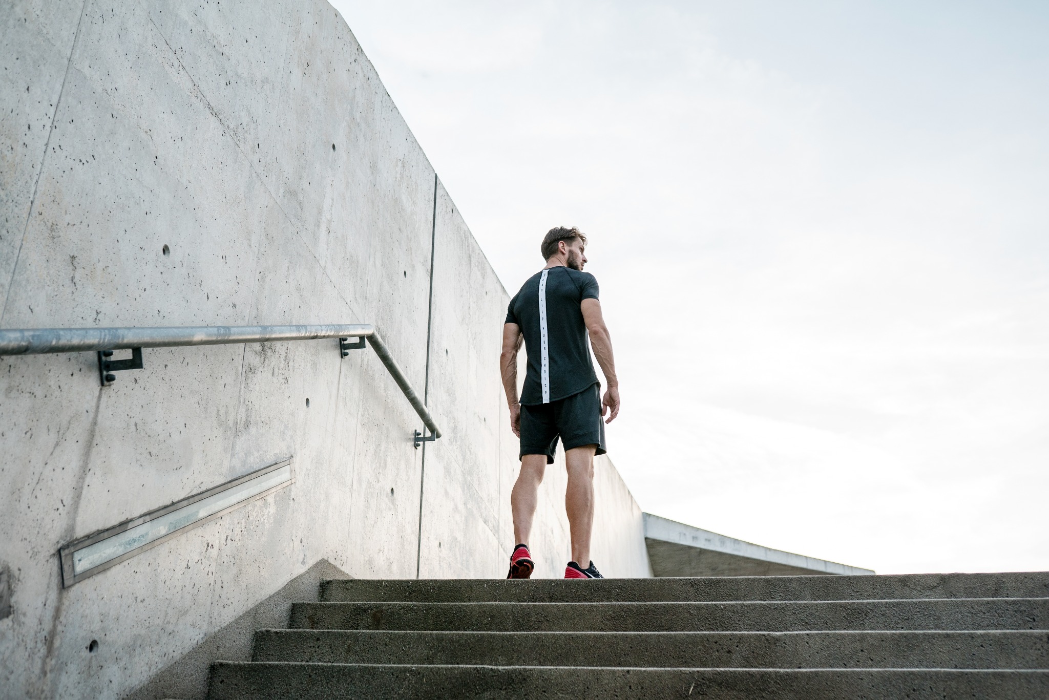 Mężczyzna w sportowym stroju wchodzi po betonowych schodach, odwrócony tyłem, z widoczną częścią betonowej ściany z metalową poręczą po lewej stronie.
