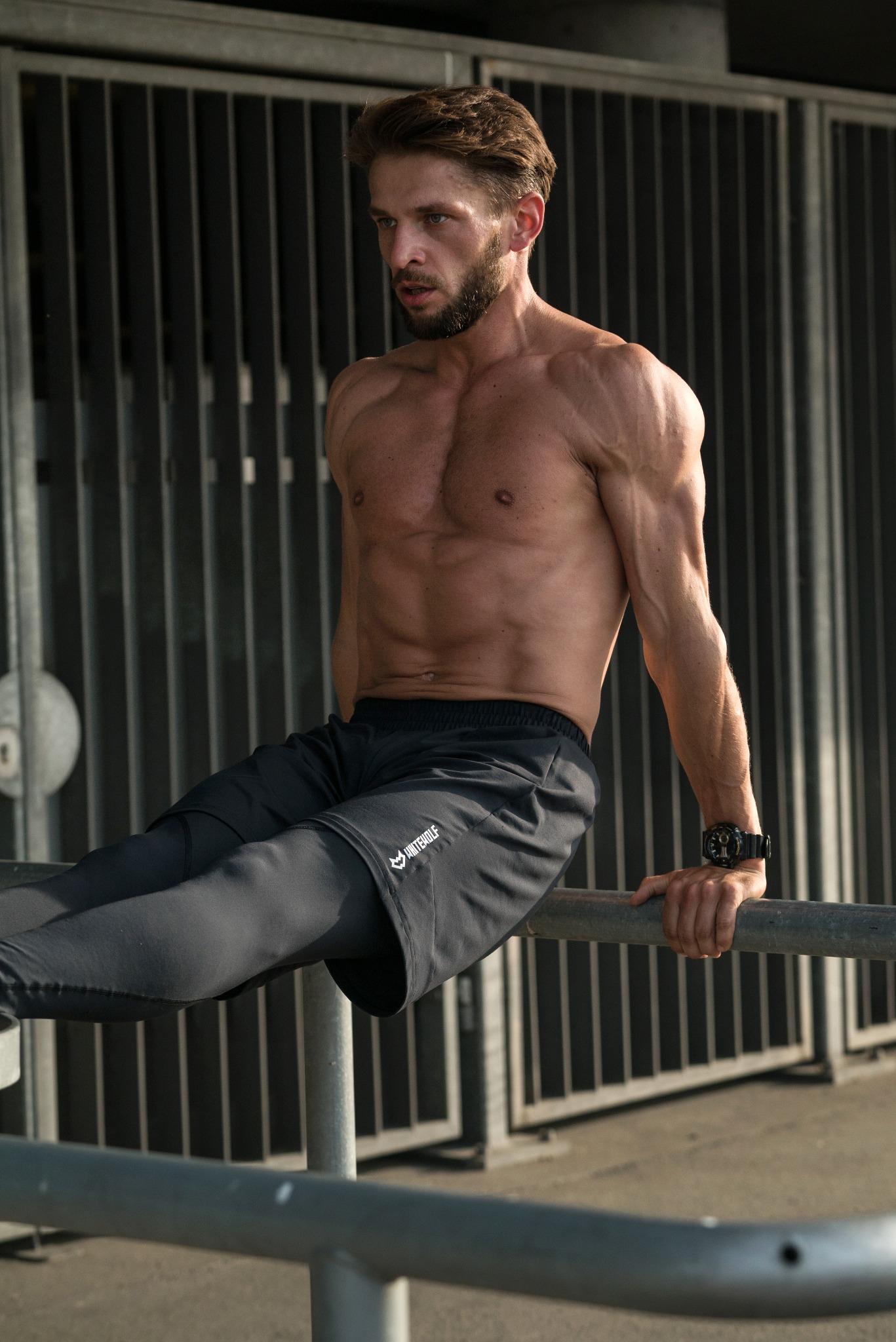 Mężczyzna bez koszulki, w sportowych spodenkach z logo, wykonuje ćwiczenie na metalowej poręczy na tle metalowego ogrodzenia.