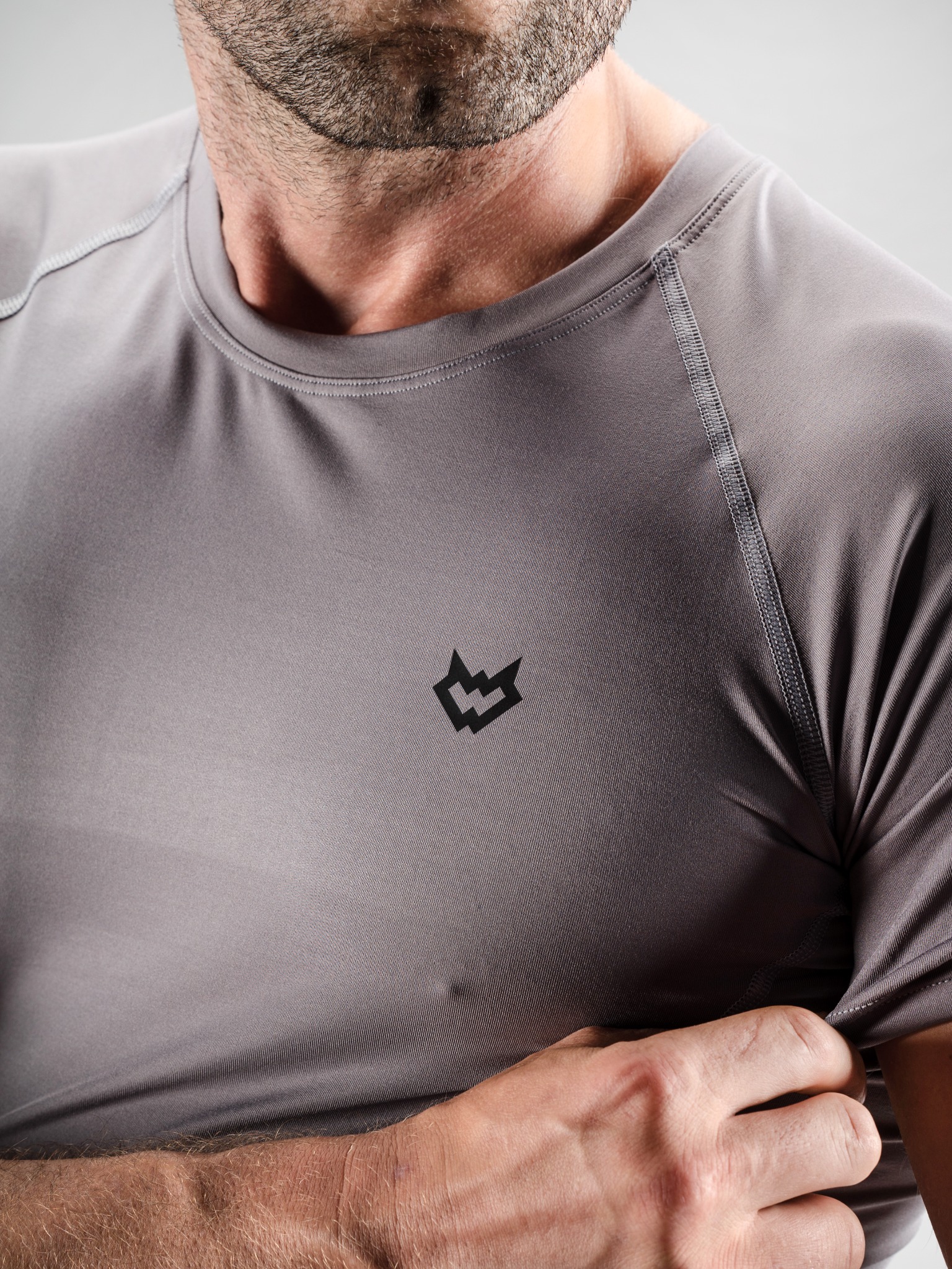 Mężczyzna w szarej, dopasowanej koszulce z krótkim rękawem i małym, czarnym logo na piersi, prezentujący ją w zbliżeniu.