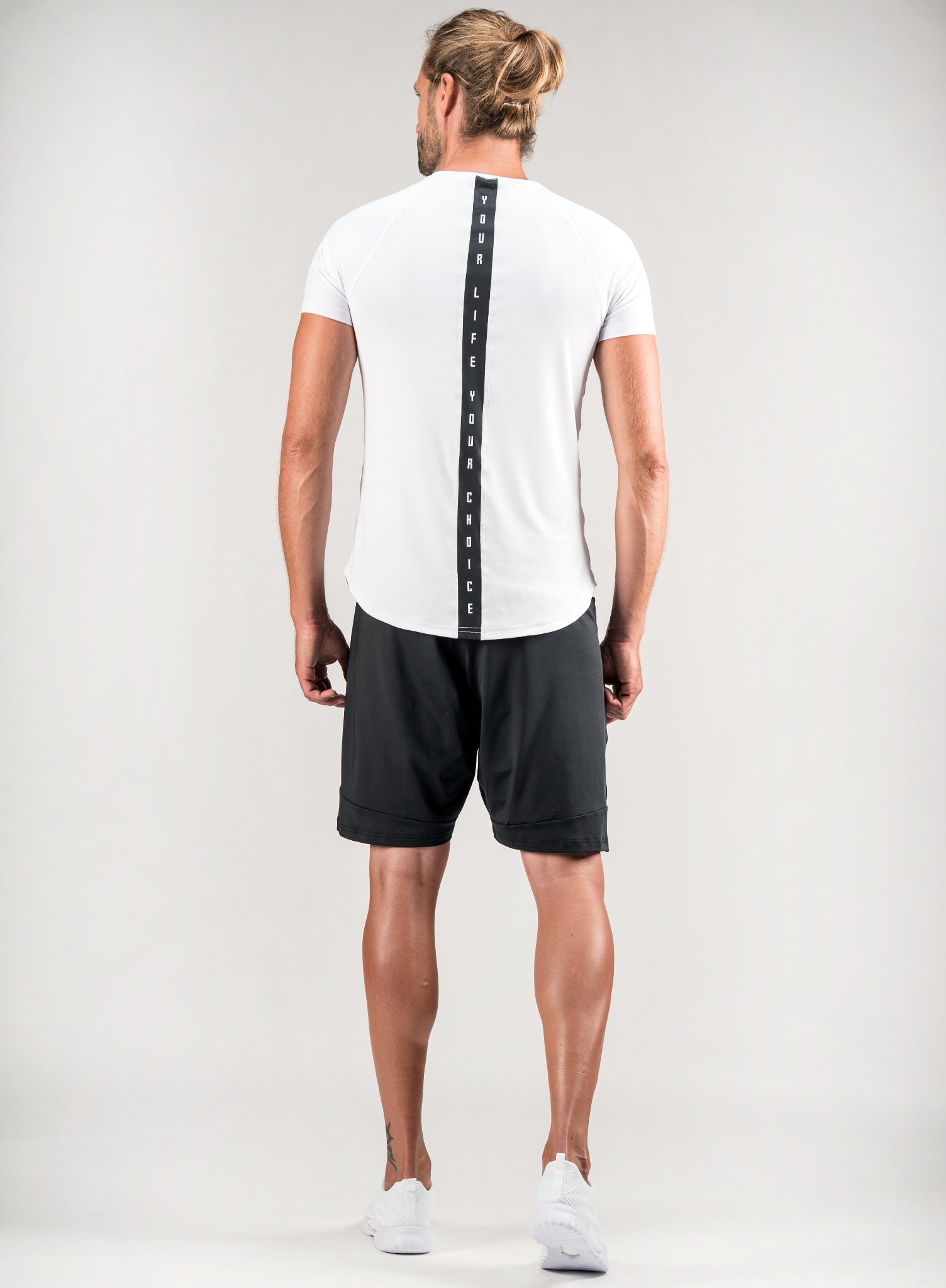 Mężczyzna w sportowym stroju, tyłem, prezentujący białą koszulkę z pionowym napisem 'YOUR LIFE YOUR CHOICE' na plecach, czarne spodenki i białe buty sportowe na jasnym tle.