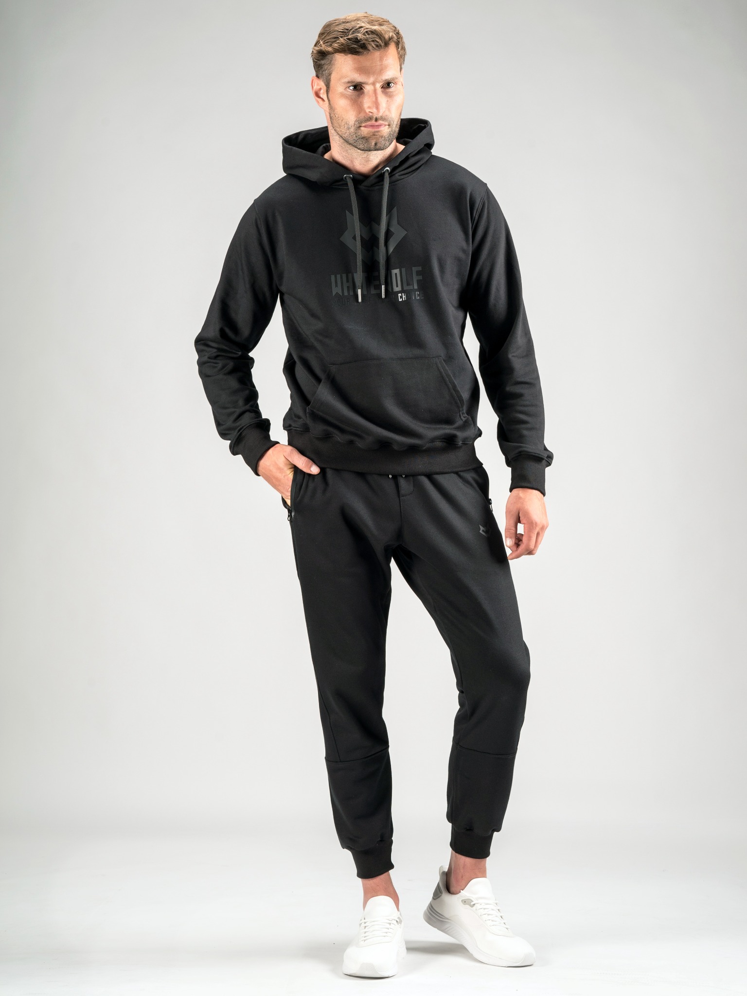 Mężczyzna w czarnym dresie z kapturem i białych butach sportowych pozuje na jasnoszarym tle.