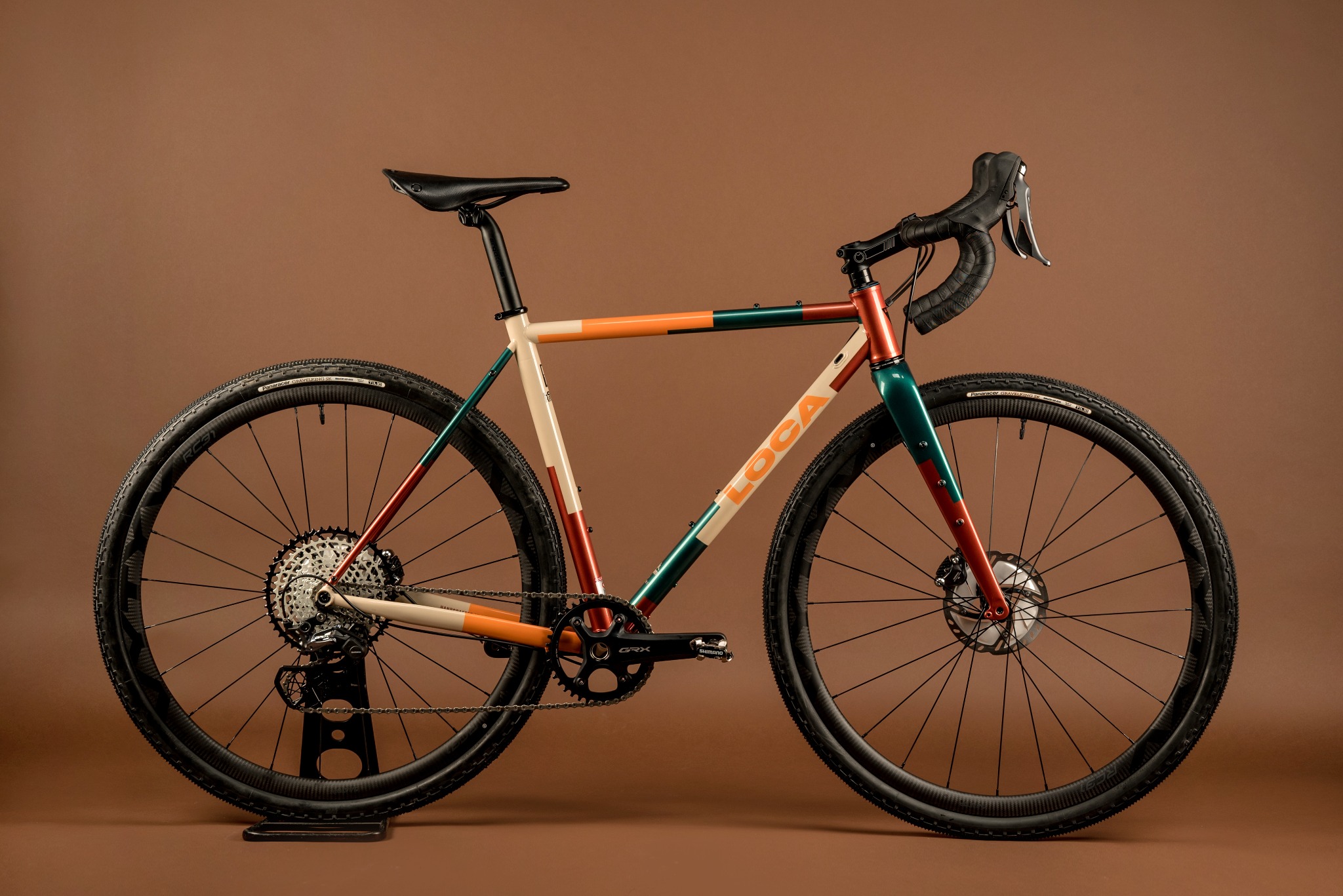 Studyjne ujęcie roweru marki Locabikes, z kremową ramą zdobioną paskami w kolorach pomarańczowym, zielonym i czerwonym, na brązowym tle.