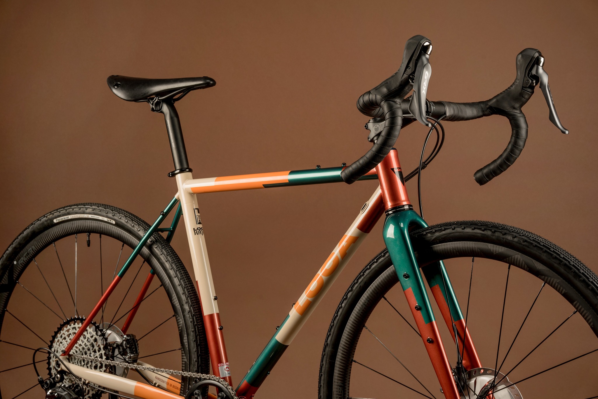Rower gravelowy z charakterystycznym malowaniem w odcieniach beżu, pomarańczy i zieleni, widoczny z boku na jednolitym, brązowym tle, eksponujący detale ramy i opon.