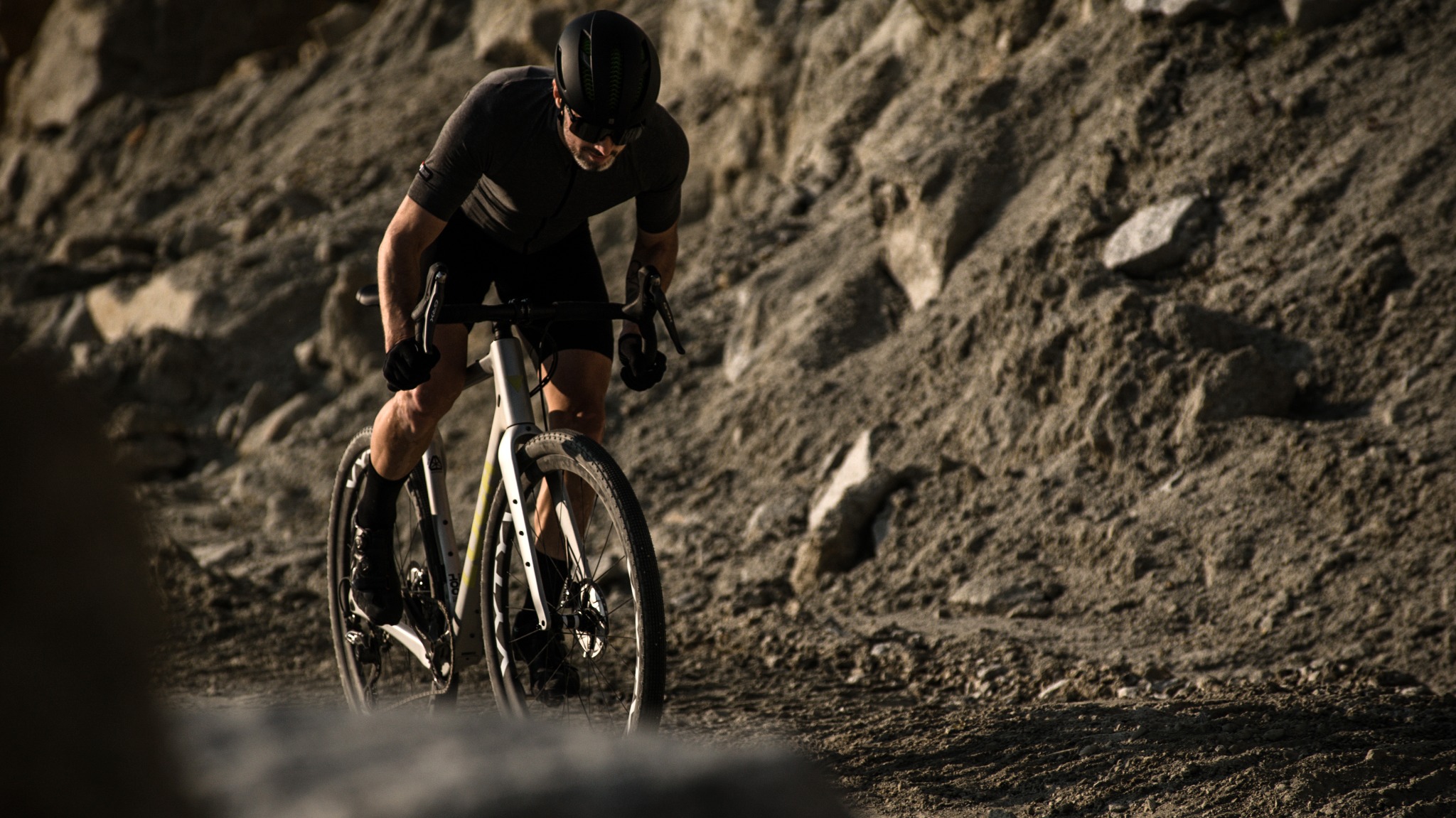 Kolarz w czarnym stroju i kasku na rowerze gravelowym, jadący po szutrowej drodze pod górę, z tłem skalistego zbocza.