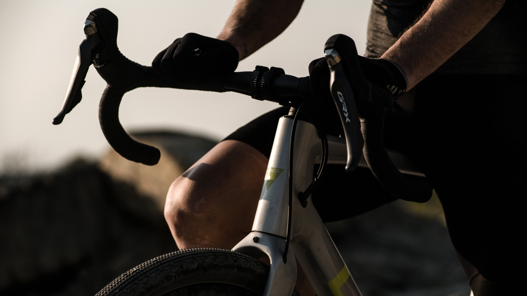 Rower gravelowy z kierownicą owiniętą czarną taśmą, widoczne manetki Shimano GRX, fragment nogi rowerzysty w czarnych spodenkach i opona z bieżnikiem szosowo-terenowym.