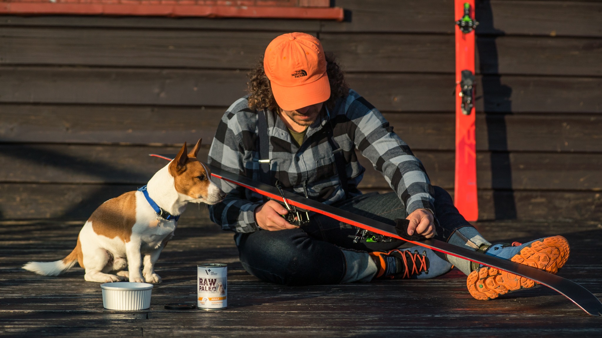 Mężczyzna w czapce i koszuli w kratę siedzi na drewnianym tarasie, regulując wiązania skiturowe; obok siedzi pies rasy Jack Russell Terrier, a w pobliżu stoi puszka karmy RawPaleo.