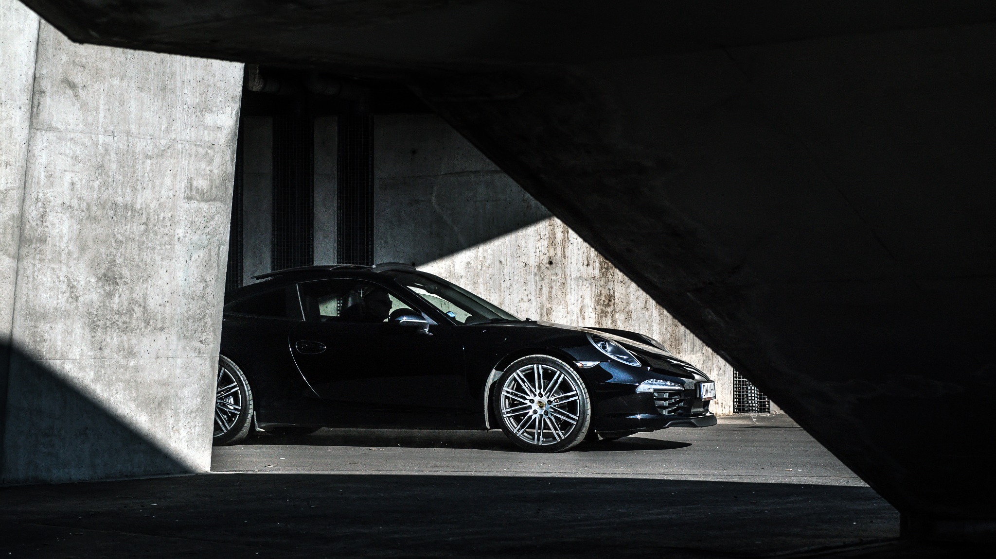 Czarny sportowy samochód Porsche z aluminiowymi felgami zaparkowany w cieniu betonowego wiaduktu, widoczne detale karoserii i oświetlenia.