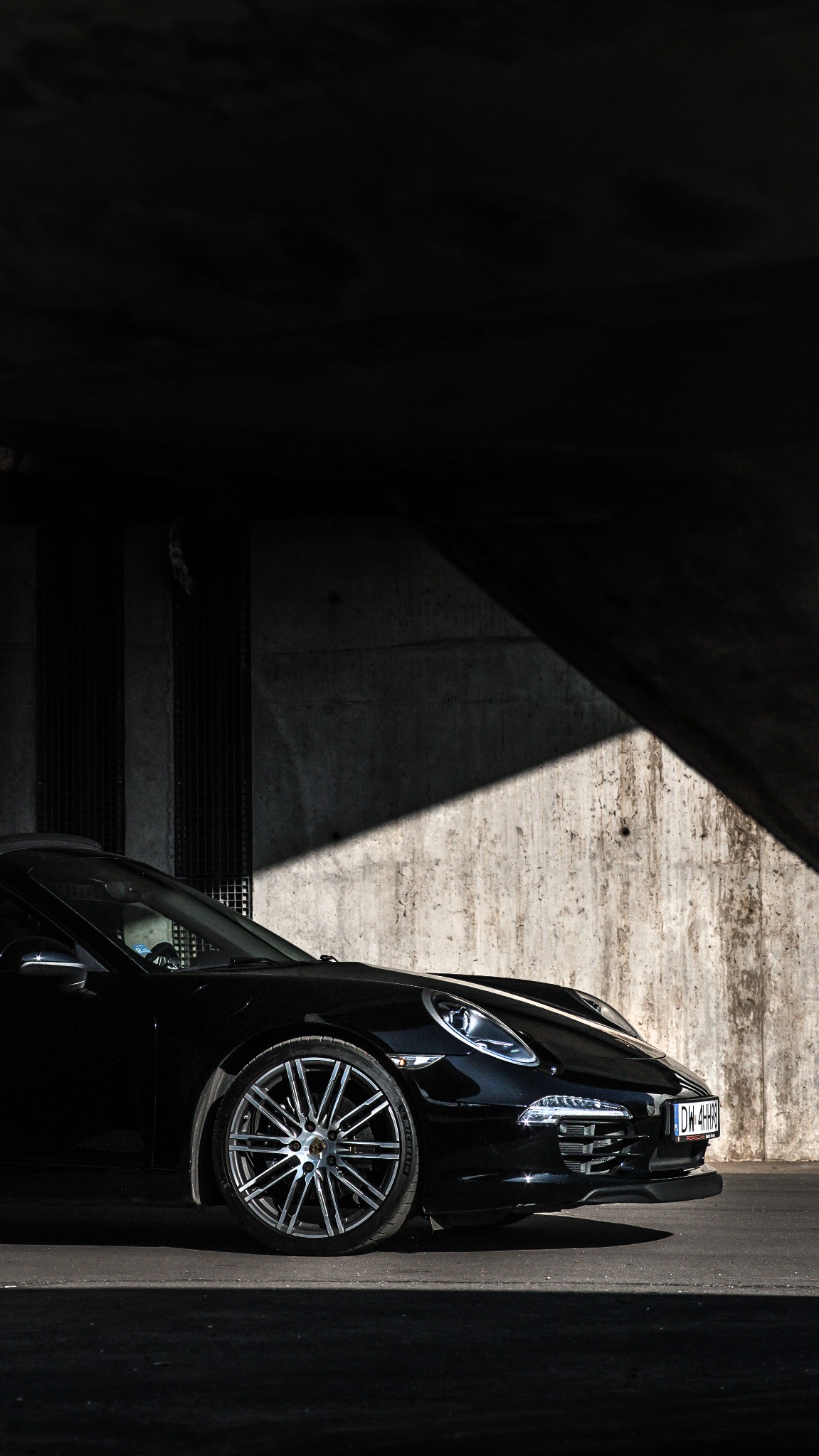 Częściowy widok czarnego samochodu sportowego Porsche z aluminiowymi felgami, zaparkowanego w zacienionym betonowym garażu z wyraźnym kontrastem światła i cienia.