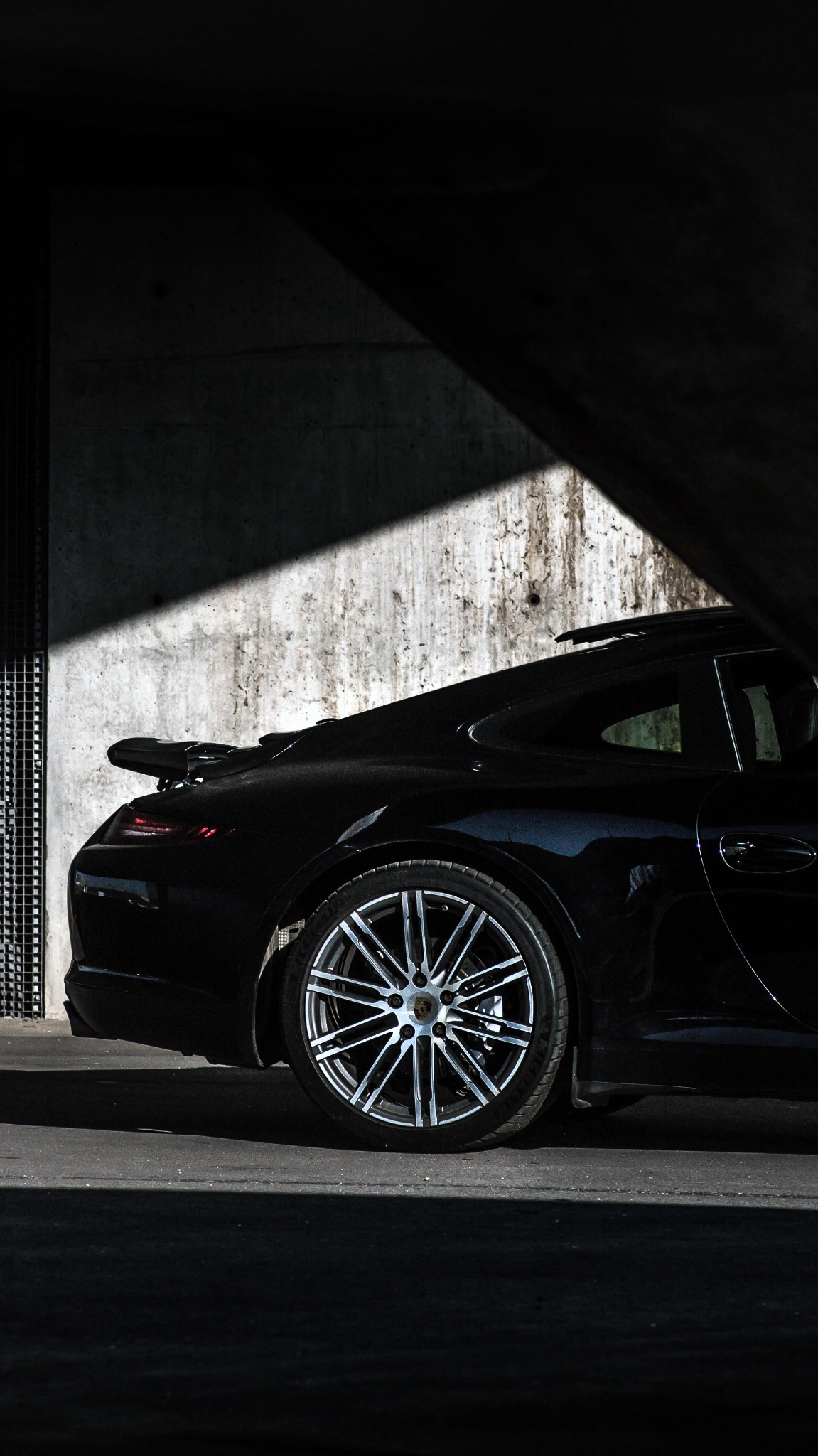 Częściowy widok czarnego Porsche 911 z aluminiowymi felgami, uchwycony w silnym kontraście światła i cienia na tle surowego, betonowego muru.