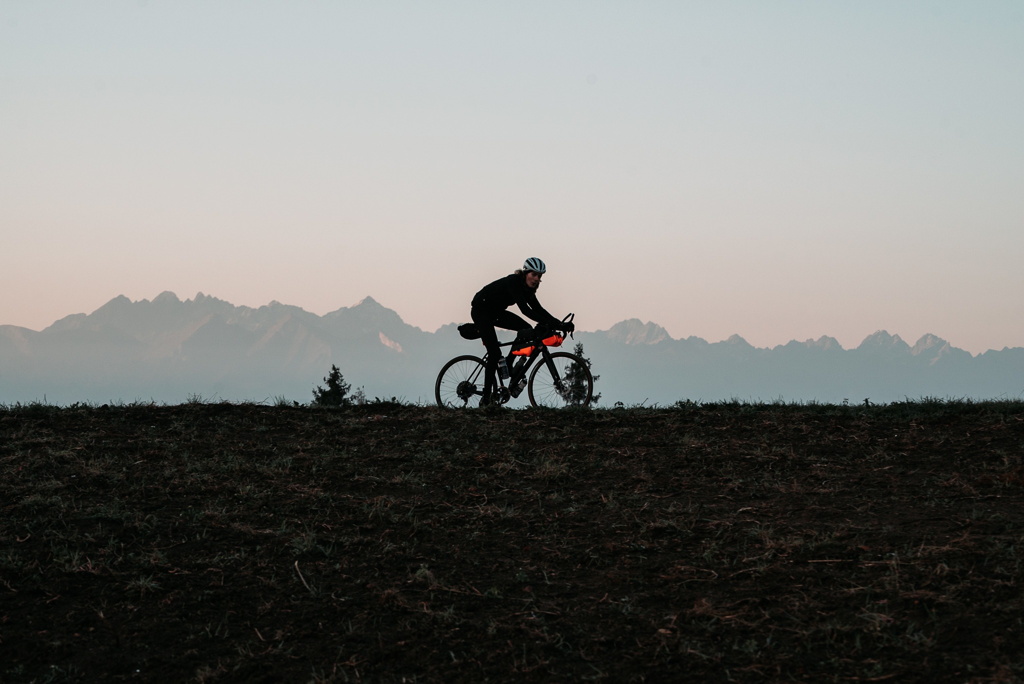 Kolarz w kasku na rowerze szosowym z torbami rowerowymi na tle pasma górskiego o zmierzchu.