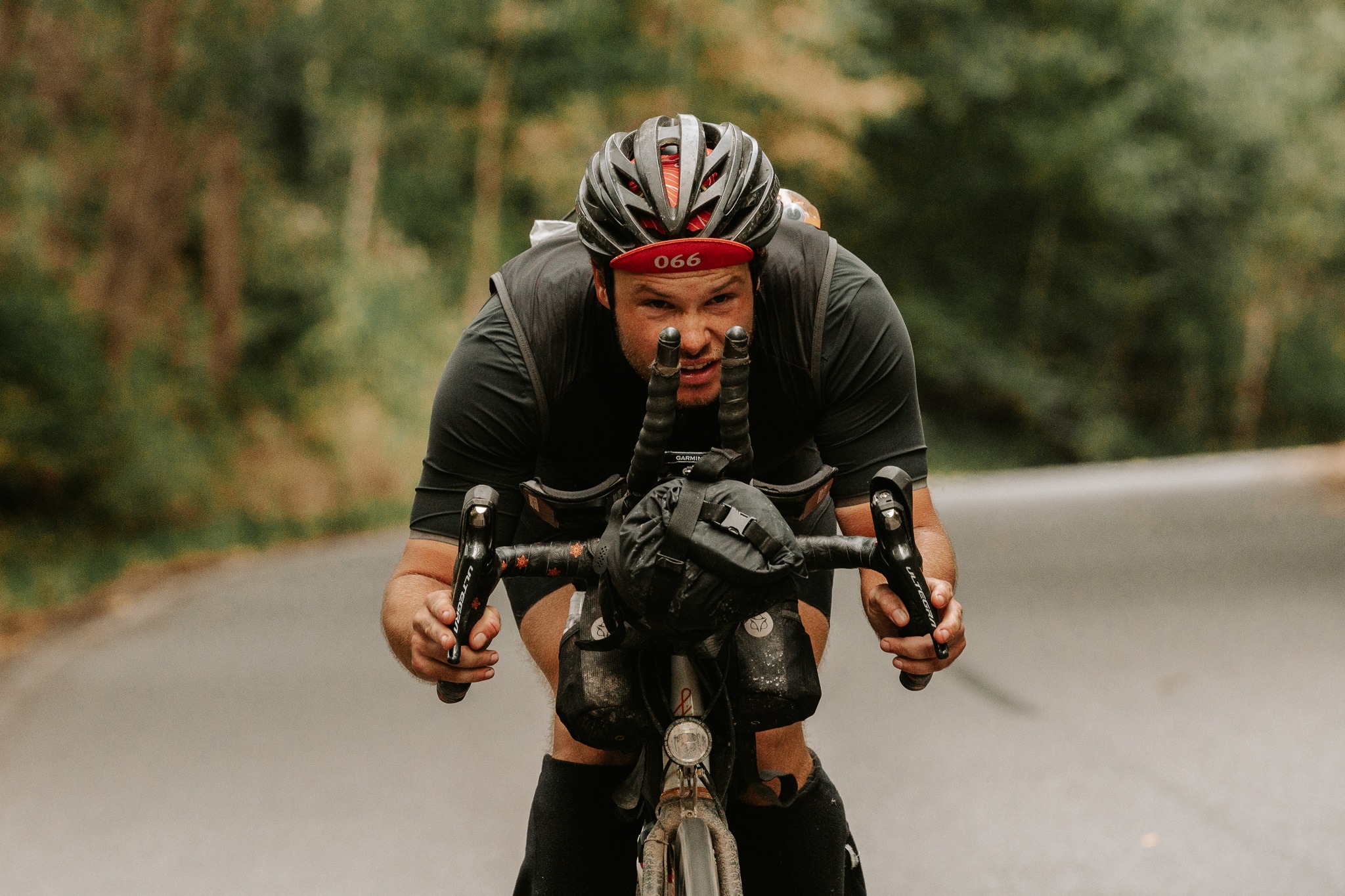 Kolarz ultra na rowerze szosowym, zdeterminowany wyraz twarzy, widoczne torby przymocowane do ramy, kask z oznaczeniem '066' na czerwonym tle, asfaltowa droga i zieleń w tle.