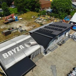 Nortmar Power Systems Centrala - Widok z góry na budynek z panelami fotowoltaicznymi na dachu, obok namiot z reklamą firmy 'Bryza' systemy rynnowe, na placu składowane materiały budowlane i zaparkowany samochód.