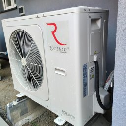 Nortmar Power Systems Centrala - Zewnętrzna jednostka pompy ciepła ROTENSO Aquami Series zamontowana na betonowych wspornikach przy szarej ścianie budynku. Widoczny wentylator, logo producenta i tabliczka znamionowa.