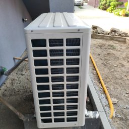 Nortmar Power Systems Centrala - Biała jednostka zewnętrzna urządzenia grzewczego, prawdopodobnie klimatyzacji lub pompy ciepła, zamontowana przy szarej ścianie budynku w trakcie prac wykończeniowych, z widoczną żółtą rurą...