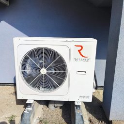 Nortmar Power Systems Centrala - Biała jednostka zewnętrzna z wentylatorem, marki Rotenso Aquami Series, zamontowana na betonowych wspornikach przy szarej ścianie budynku. Widoczne elementy instalacji i otoczenie z piaskiem...