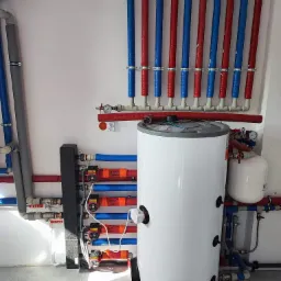 Biały zasobnik ciepłej wody z podłączonymi rurami w kolorach czerwonym i niebieskim, zabezpieczonymi izolacją, zamontowany w pomieszczeniu z szarą podłogą i białymi ścianami. Widoczne pompy...