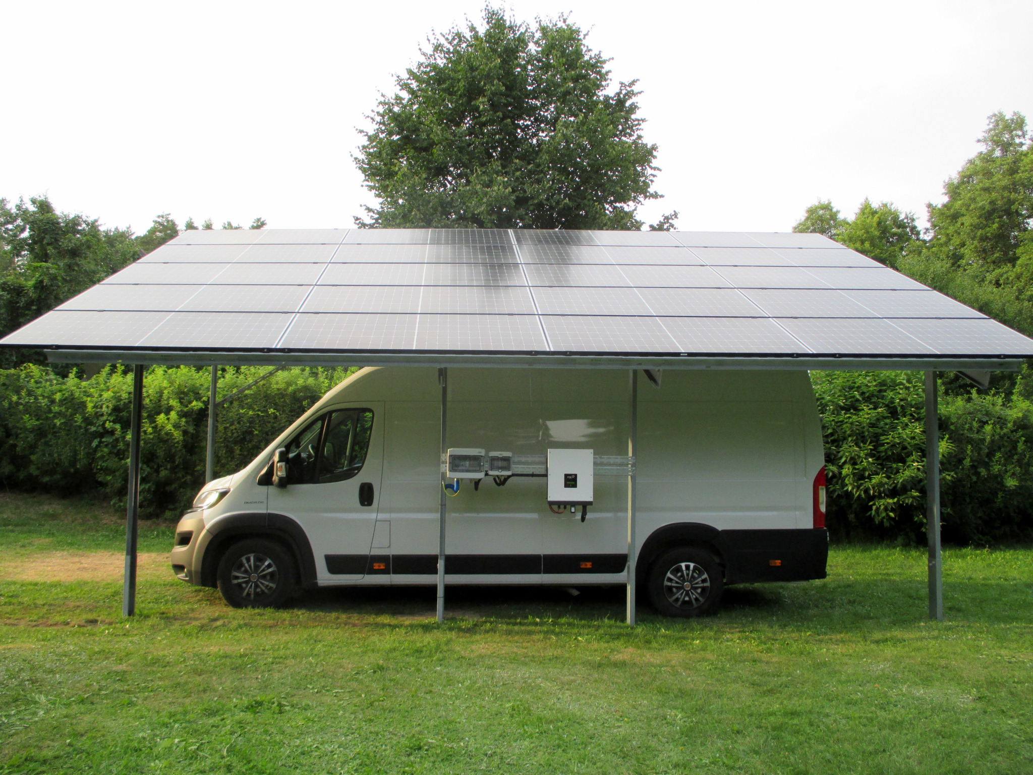 Biały samochód dostawczy zaparkowany pod zadaszeniem z paneli fotowoltaicznych na metalowej konstrukcji, z widocznym inwerterem i skrzynką przyłączeniową na boku pojazdu, na tle zielonego trawnika...