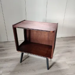 Ręcznie wykonana, drewniana szafka nocna w stylu retro z ciemnymi nóżkami, ustawiona na panelach podłogowych w jasnym odcieniu drewna, na tle jasnoszarej ściany.