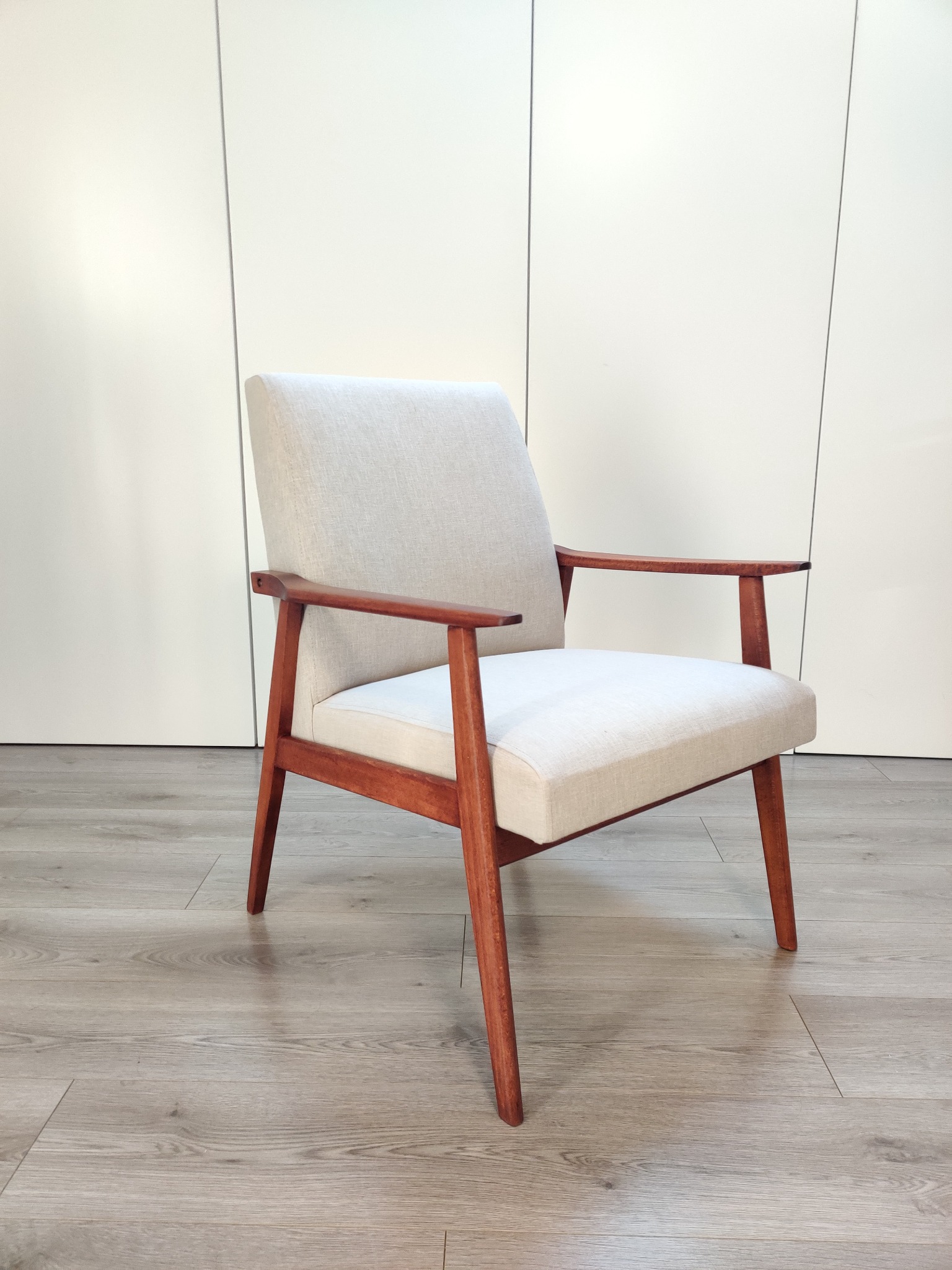 Odrestaurowany fotel w stylu retro z drewnianymi podłokietnikami i jasną tapicerką na tle białej ściany z paneli.