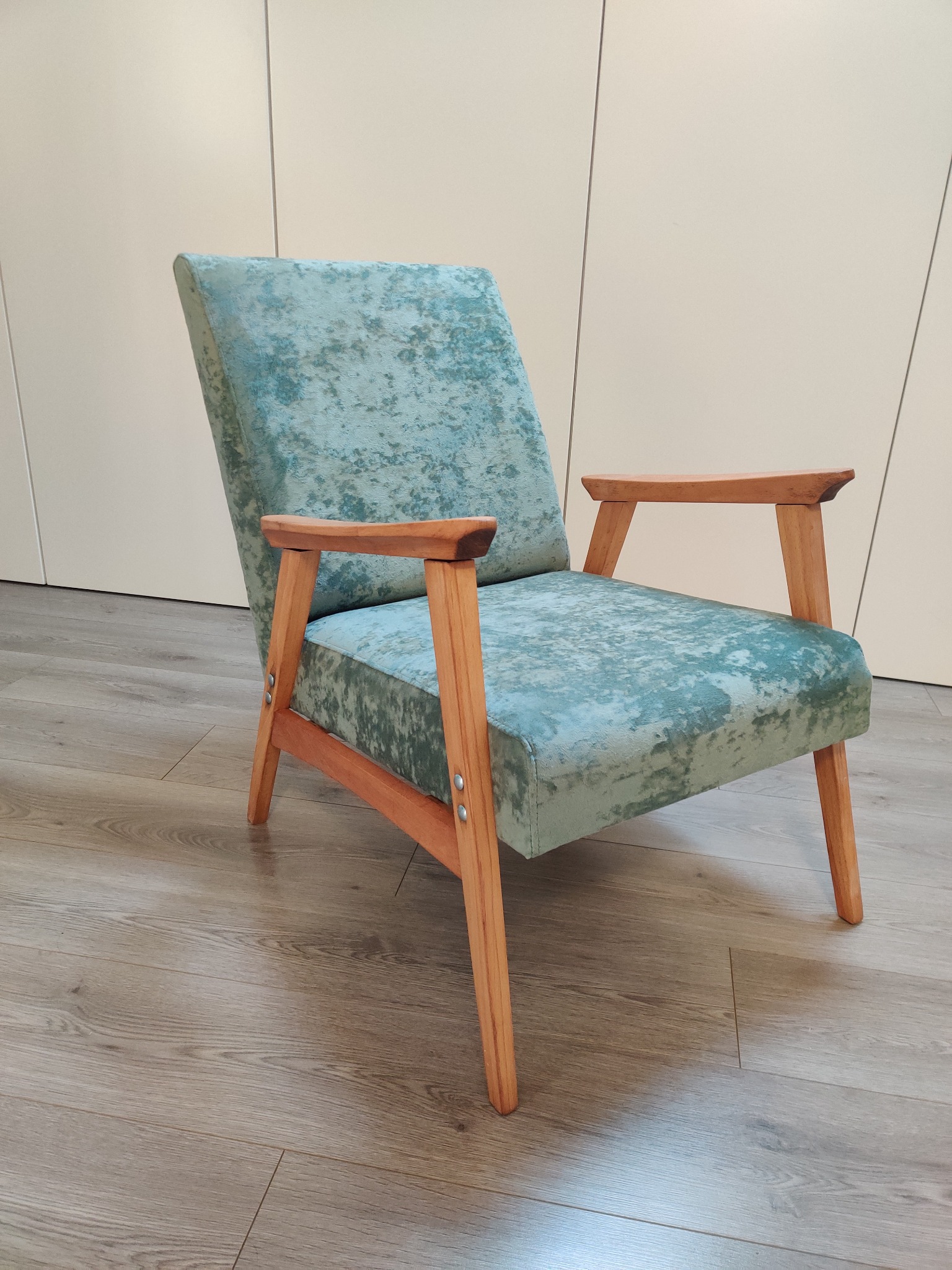 Odnowiony fotel w stylu vintage z turkusową tapicerką o aksamitnej fakturze i drewnianymi podłokietnikami, stojący na panelach podłogowych na tle białej szafy.