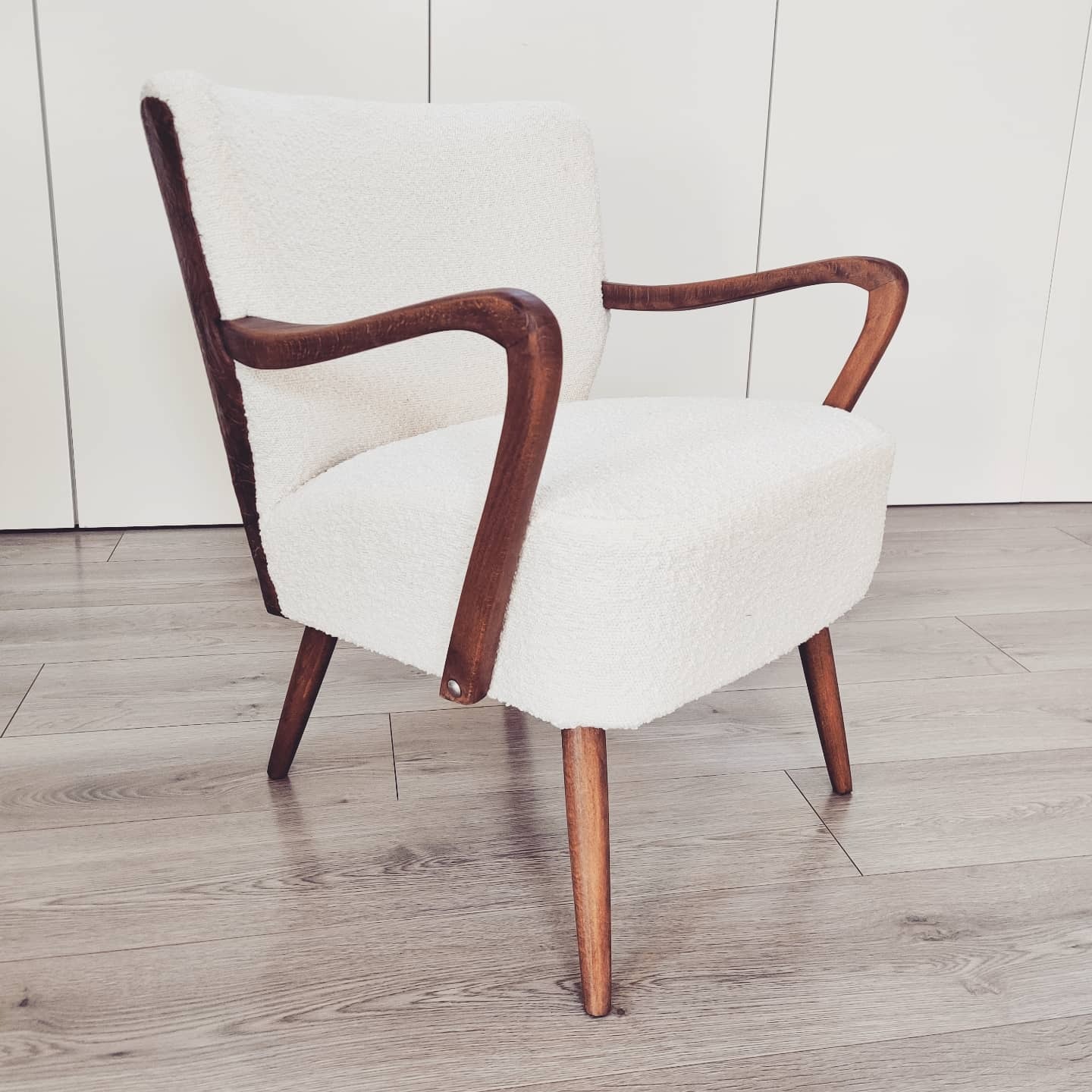 Fotel w stylu retro z białym, teksturowanym obiciem i drewnianymi nogami oraz podłokietnikami, ustawiony na jasnej podłodze z paneli.