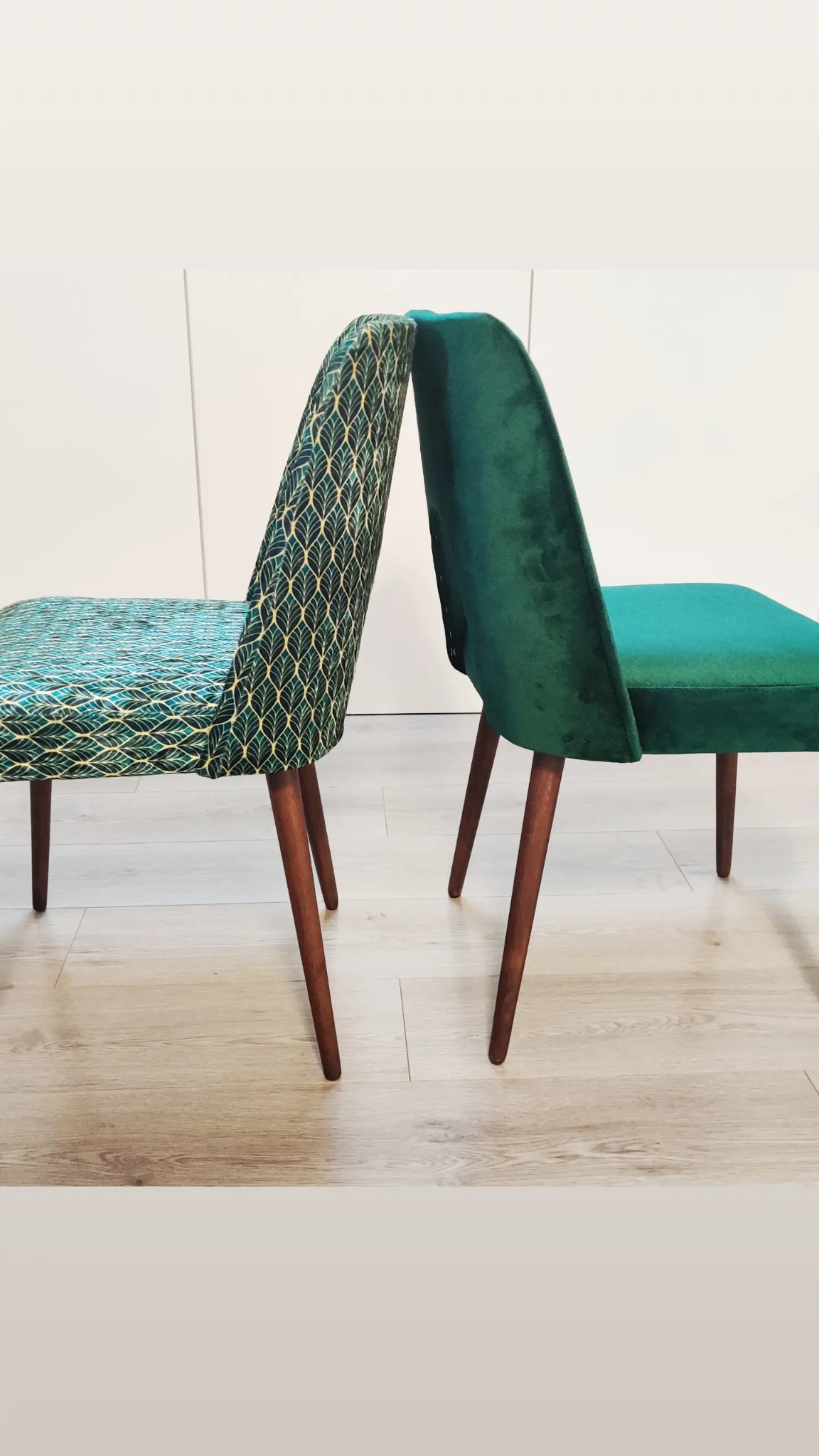 Dwa krzesła z drewnianymi nogami: jedno z zielonym wzorem liści na jasnym tle, drugie obite ciemnozielonym welurem, ustawione tyłem do siebie na jasnej podłodze.