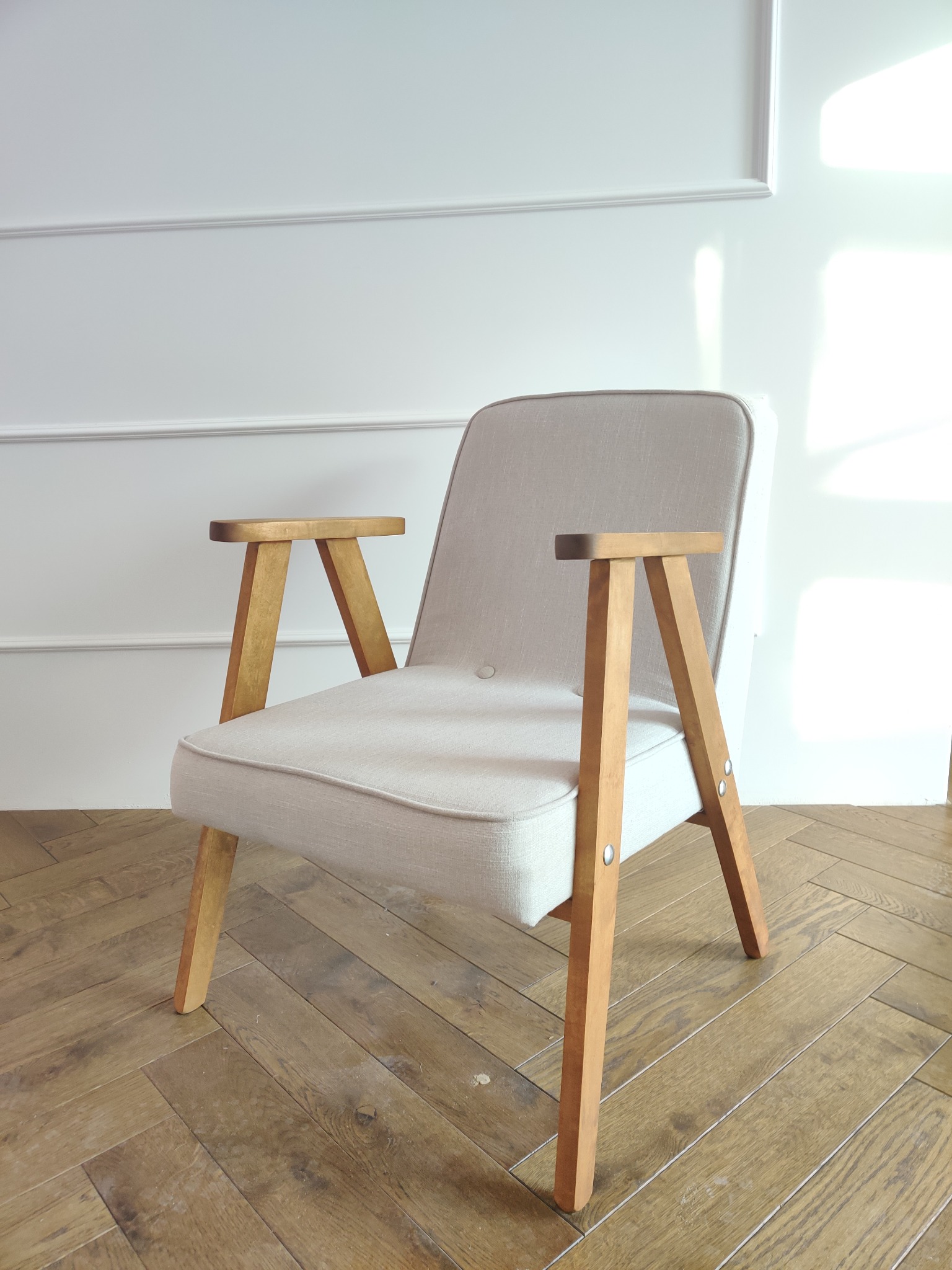 Beżowy fotel w stylu retro z drewnianymi nogami i podłokietnikami, ustawiony na parkiecie w jodełkę, na tle białej ściany z dekoracyjną listwą.