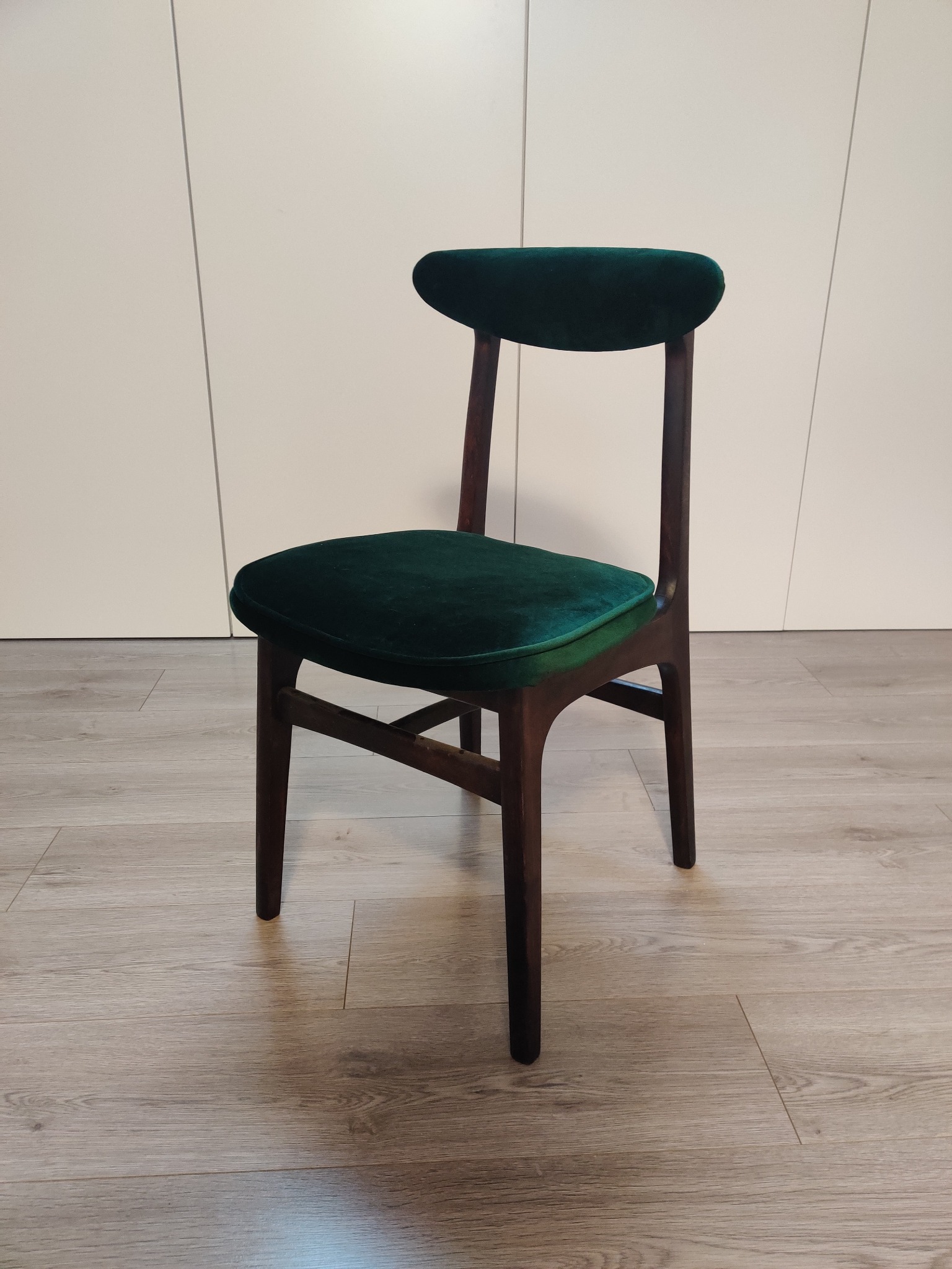 Krzesło z ciemnego drewna z nową, ciemnozieloną, aksamitną tapicerką siedziska i oparcia, stojące na panelach podłogowych na tle białej szafy.