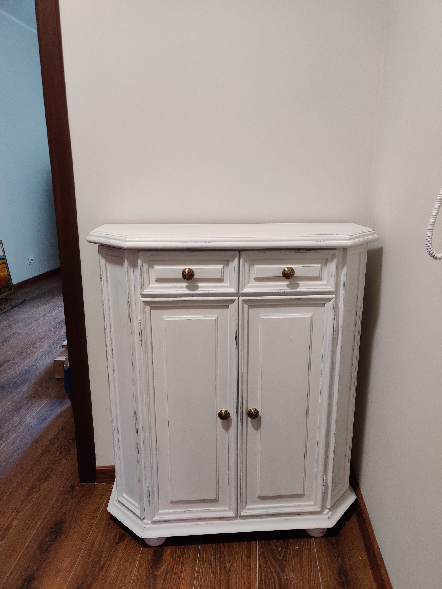 Biała, postarzana szafka narożna z dwoma szufladami i drzwiczkami, z okrągłymi, złotymi gałkami, na tle drewnianej podłogi i jasnej ściany.