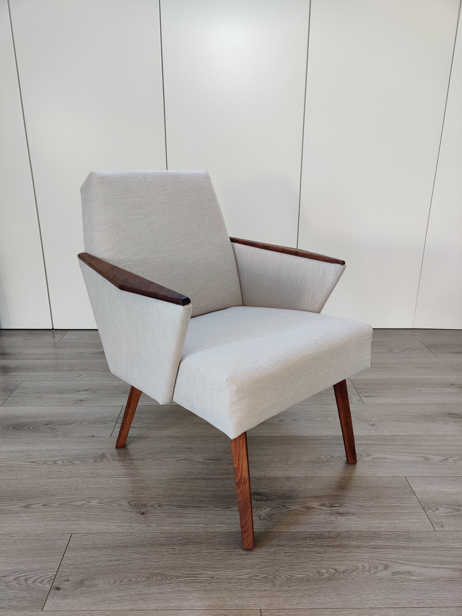 Beżowy fotel w stylu mid-century modern z drewnianymi podłokietnikami i nogami, ustawiony na panelach podłogowych na tle białej ściany z pionowymi panelami.