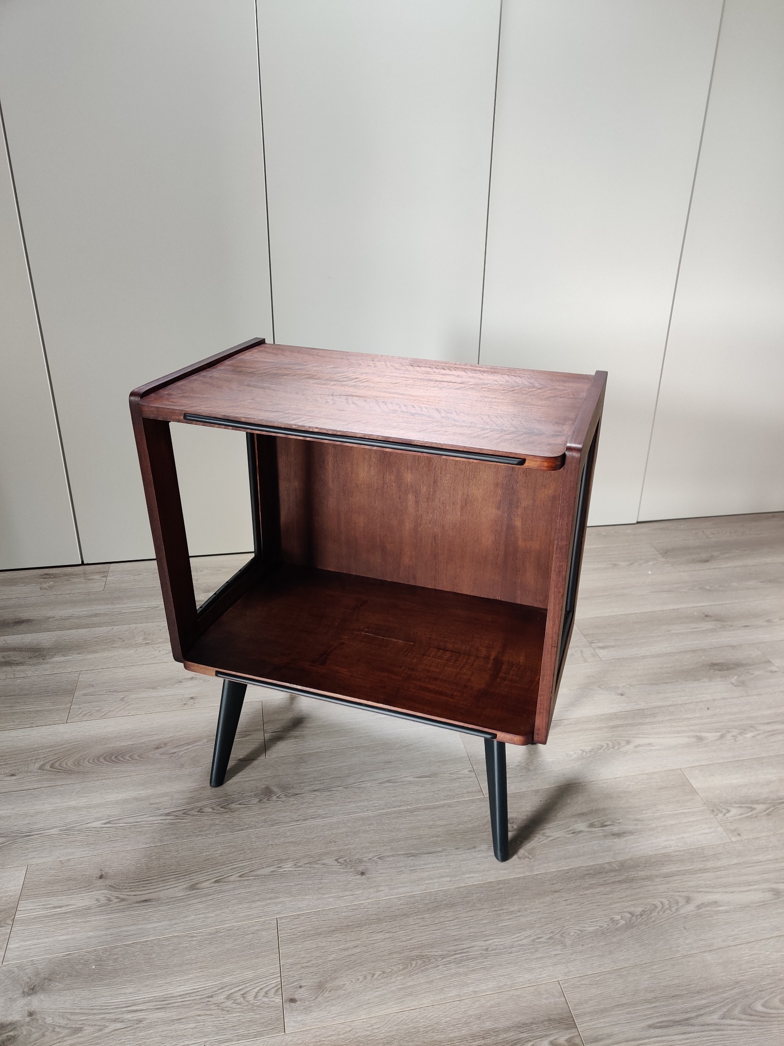 Ręcznie wykonana, drewniana szafka nocna w stylu retro z ciemnymi nóżkami, ustawiona na panelach podłogowych w jasnym odcieniu drewna, na tle jasnoszarej ściany.